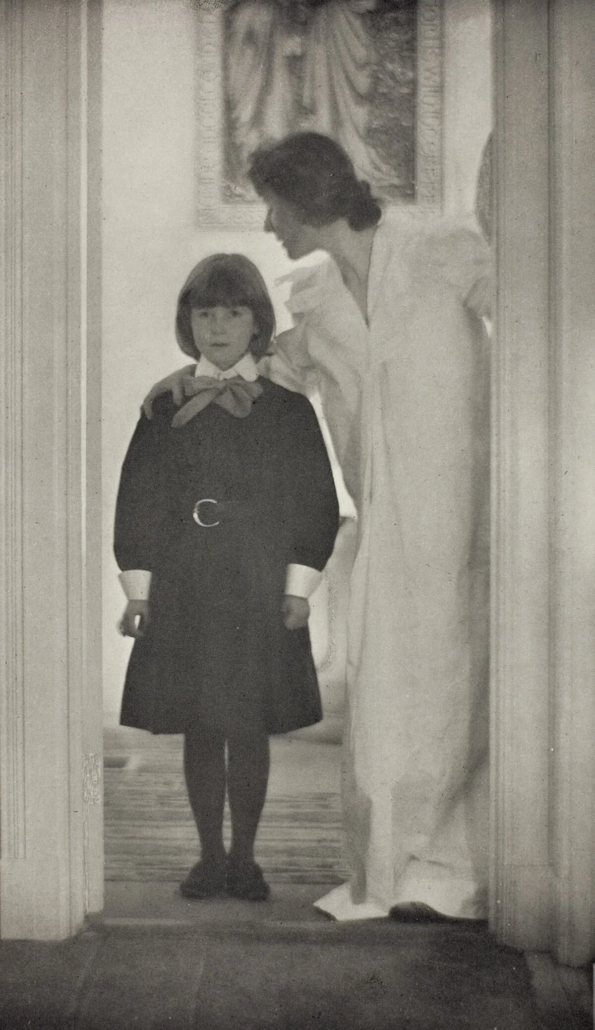 Photograph of a young girl in a doorwary wearing a black dress and a woman in a long, draping garment with her arm around her.