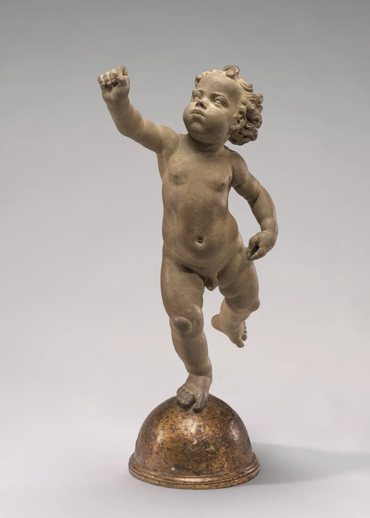 Sculpted in clay, a toddler balances his plump body on his right foot as he raises his left leg and right arm.