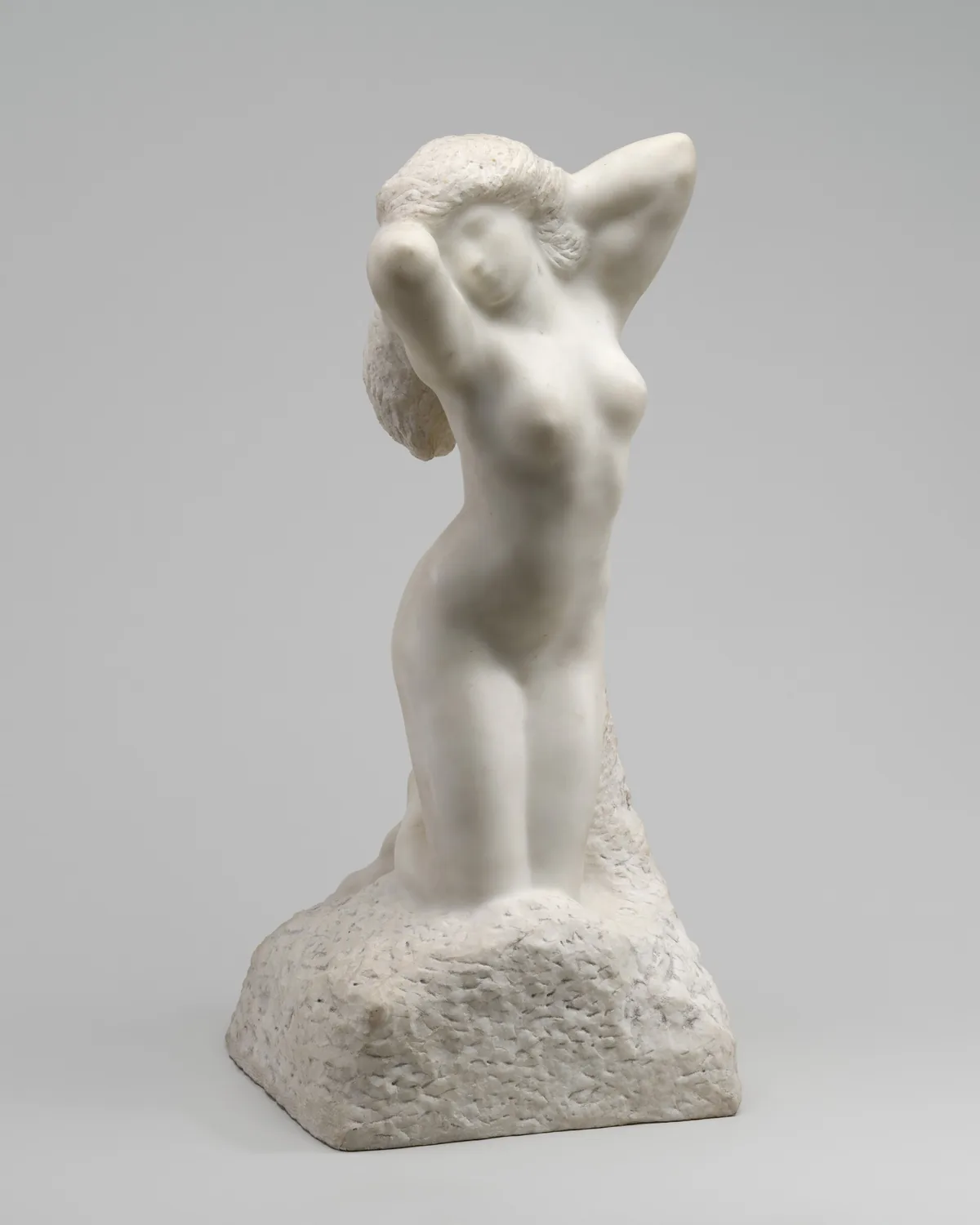 A woman kneeling with both arms raised by her head is carved from white marble in this freestanding sculpture.