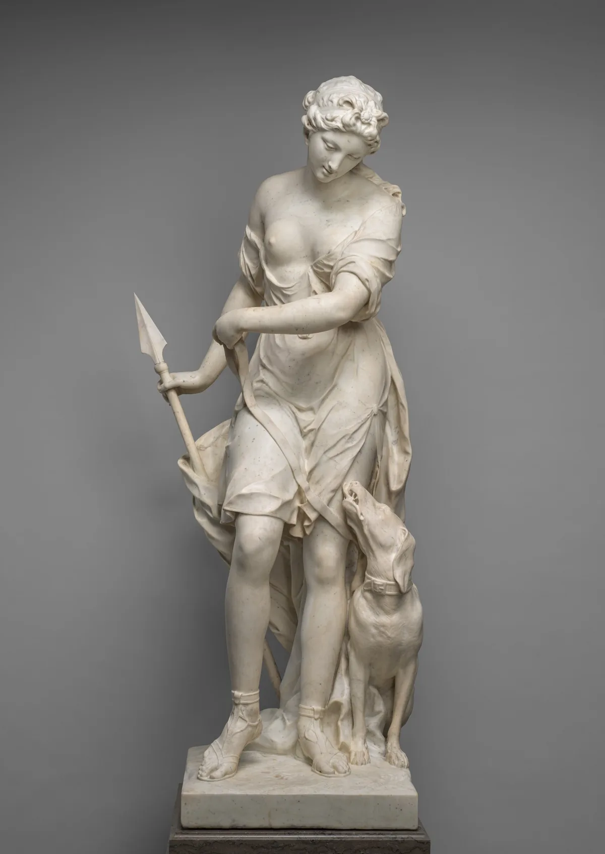 Carved from white marble, a young woman holds a short spear by one side and looks down toward a dog standing by her other side in this free-standing sculpture.