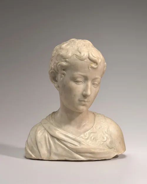 The head and shoulders of a young boy with short, curly hair is carved from cream-white marble in this free-standing sculpture.