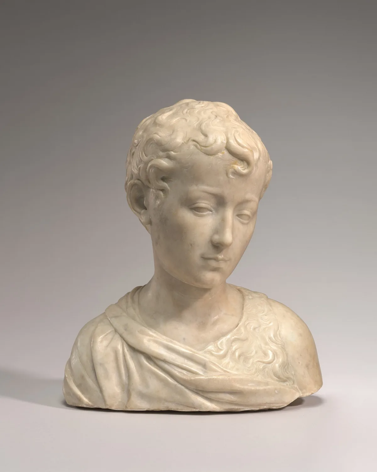 The head and shoulders of a young boy with short, curly hair is carved from cream-white marble in this free-standing sculpture.