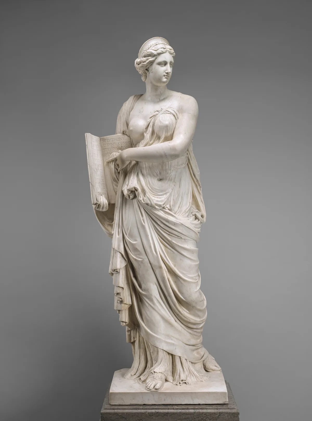 A woman carved from cream-white marble stands holding an open book to which she points in this free-standing sculpture.