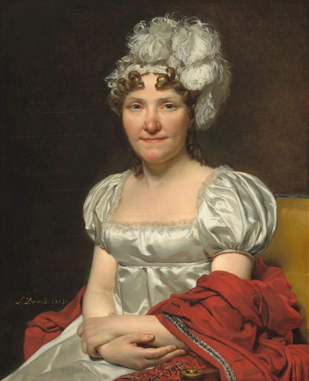 Shown from the lap up, a woman with peachy skin, wearing a white silk dress and feathered headpiece, sits facing and looking at us in a harvest-yellow, upholstered chair in this vertical portrait painting.