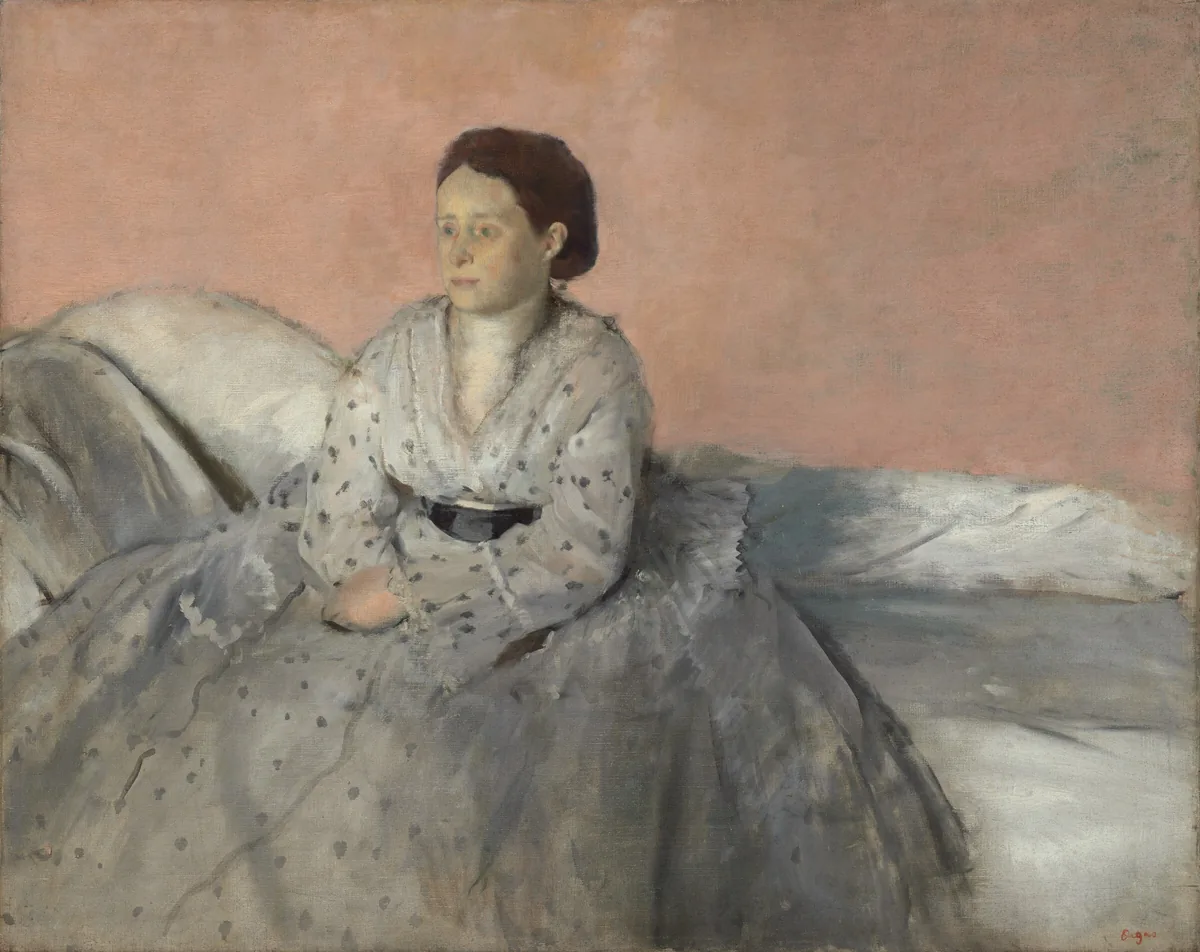 Shown from about the ankles up, a woman with ashen, pale skin, wearing a full, silvery-white dress with gray dots, sits on a low-backed white sofa against a pale peach background in this horizontal portrait painting.