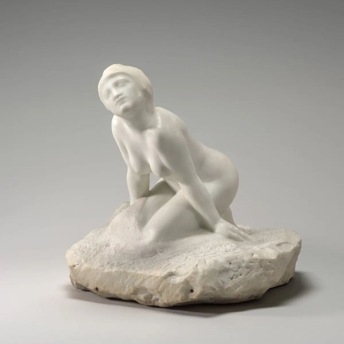 The sculpture depicts a nude female figure kneeling and leaning forward on a textured base resembling a natural, rocky surface.