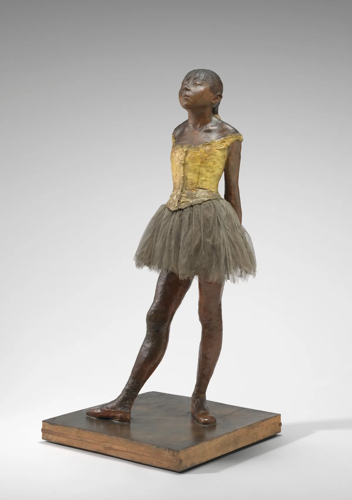Sculpted with rich brown wax, a young ballerina stands with her arms straight, hands clasped behind her back, and one foot in front of the other on a square wooden base.