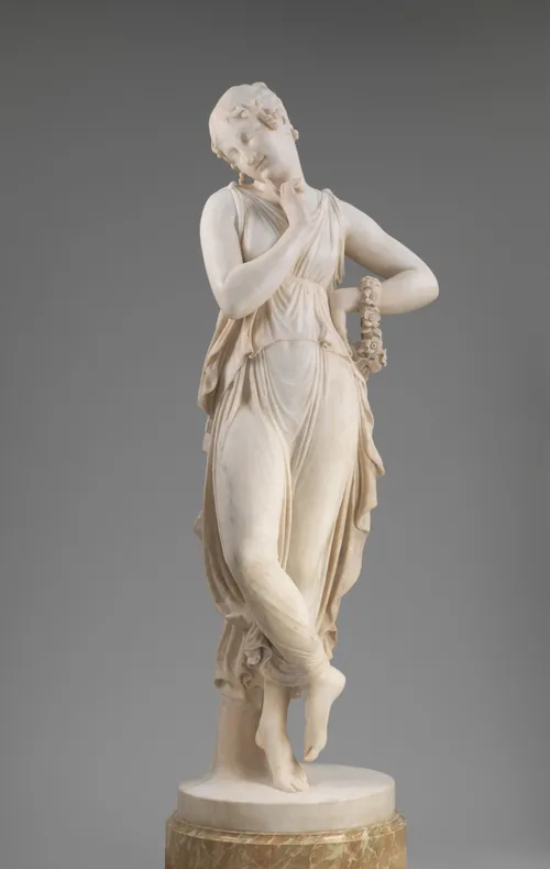 Carved from white marble, a woman balances on the ball of one foot as she holds one finger to her chin in this freestanding sculpture.