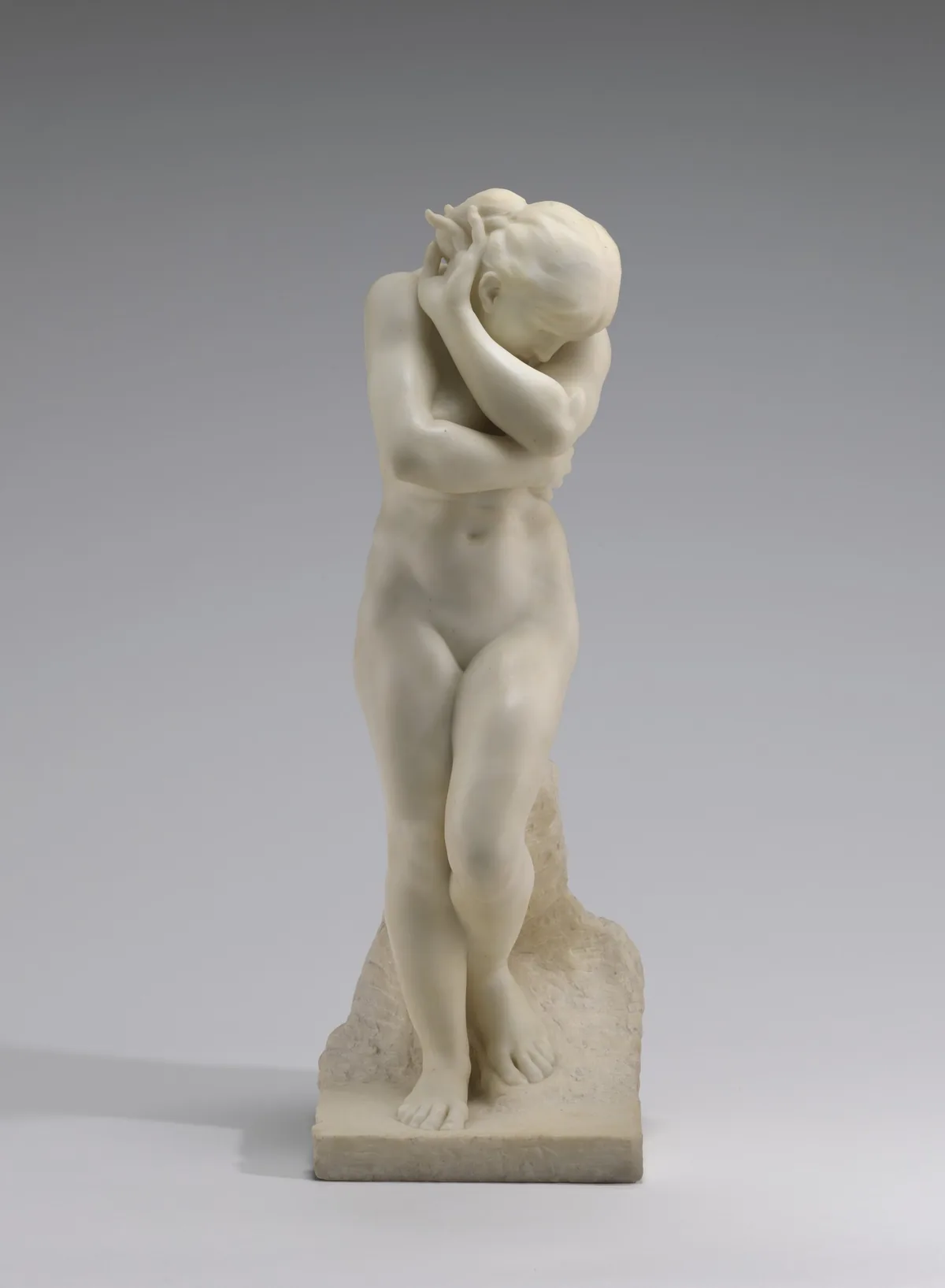 The sculpture depicts a nude woman standing with her arms wrapped around her body.