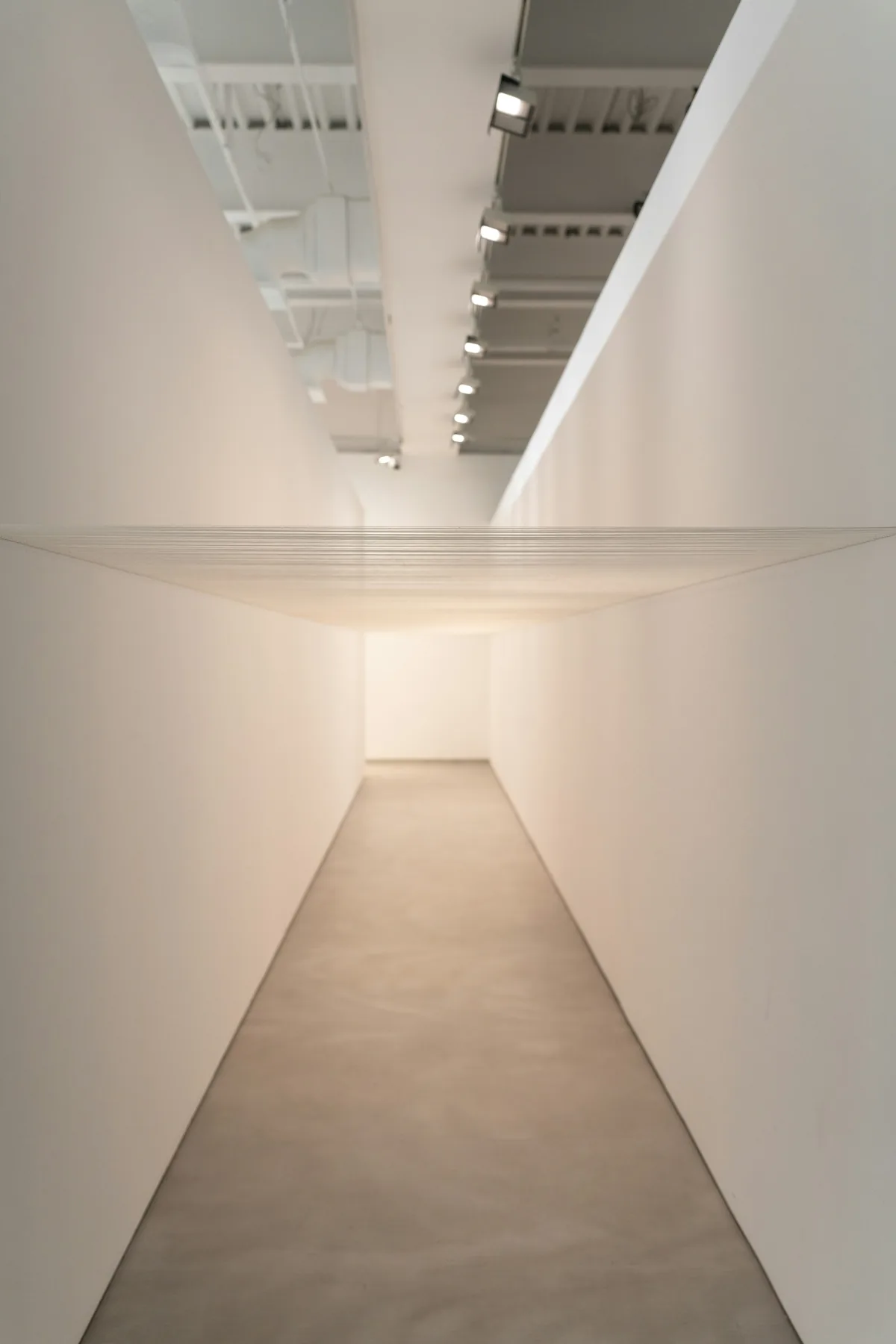 A minimalist white corridor leading toward a suspended thread installation.