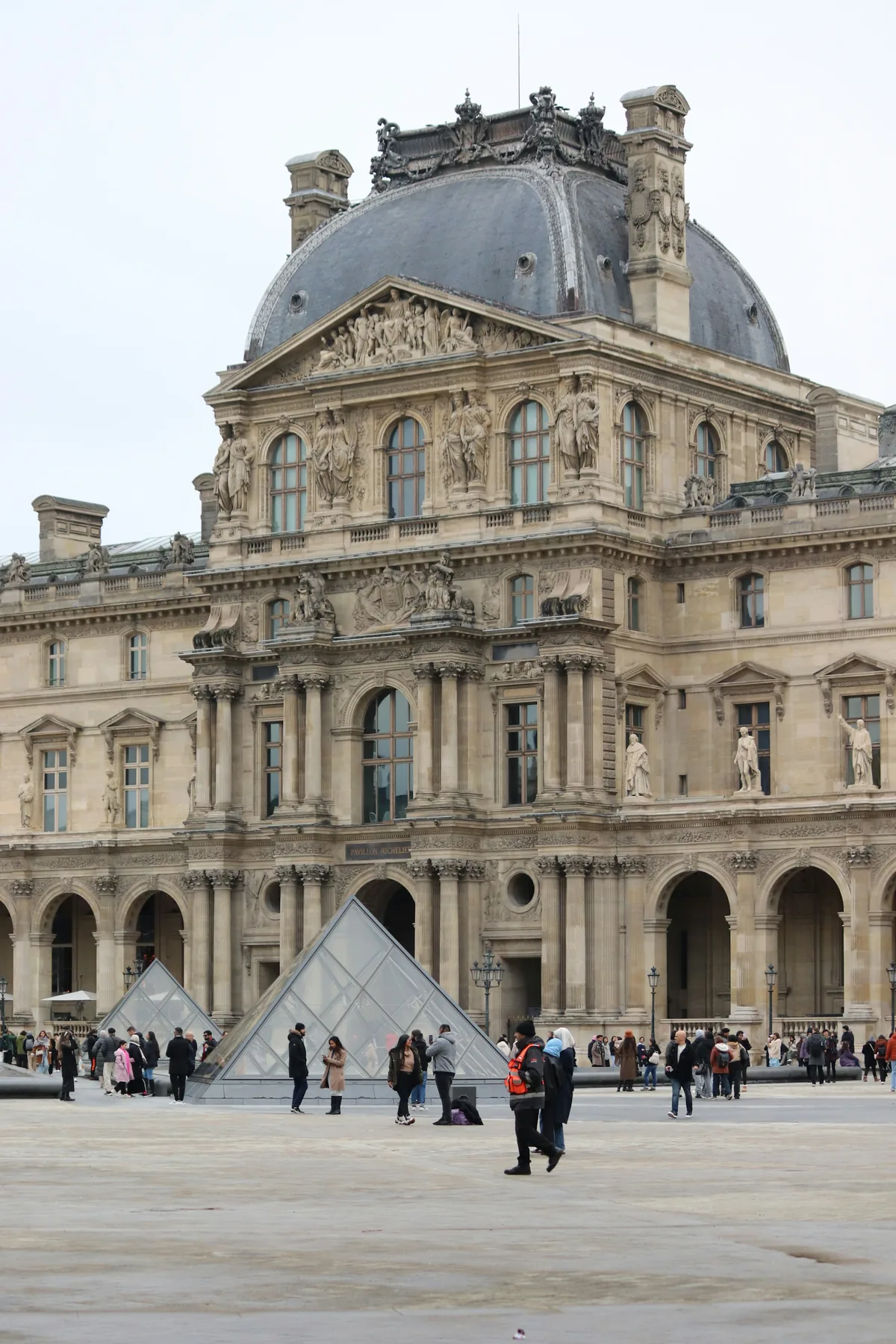 The Richelieu wing of the Louvre behind Pei’s glass pyramid, visitors crossing the Cour Napoléon.