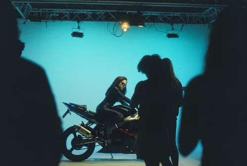 A woman in leather poses on a motorcycle under blue studio light, shot backstage during a music video shoot.