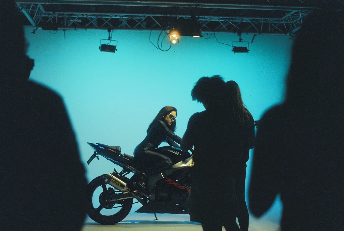 A woman in leather poses on a motorcycle under blue studio light, shot backstage during a music video shoot.