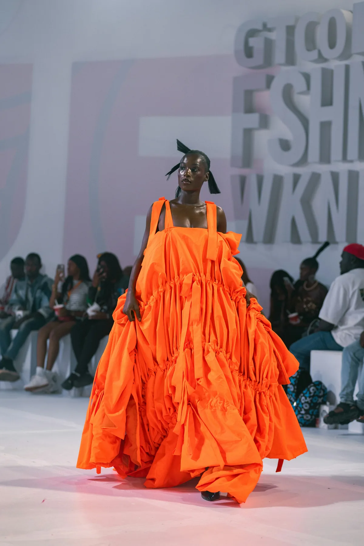 A model in a voluminous orange gown walks a runway at a fashion week show.