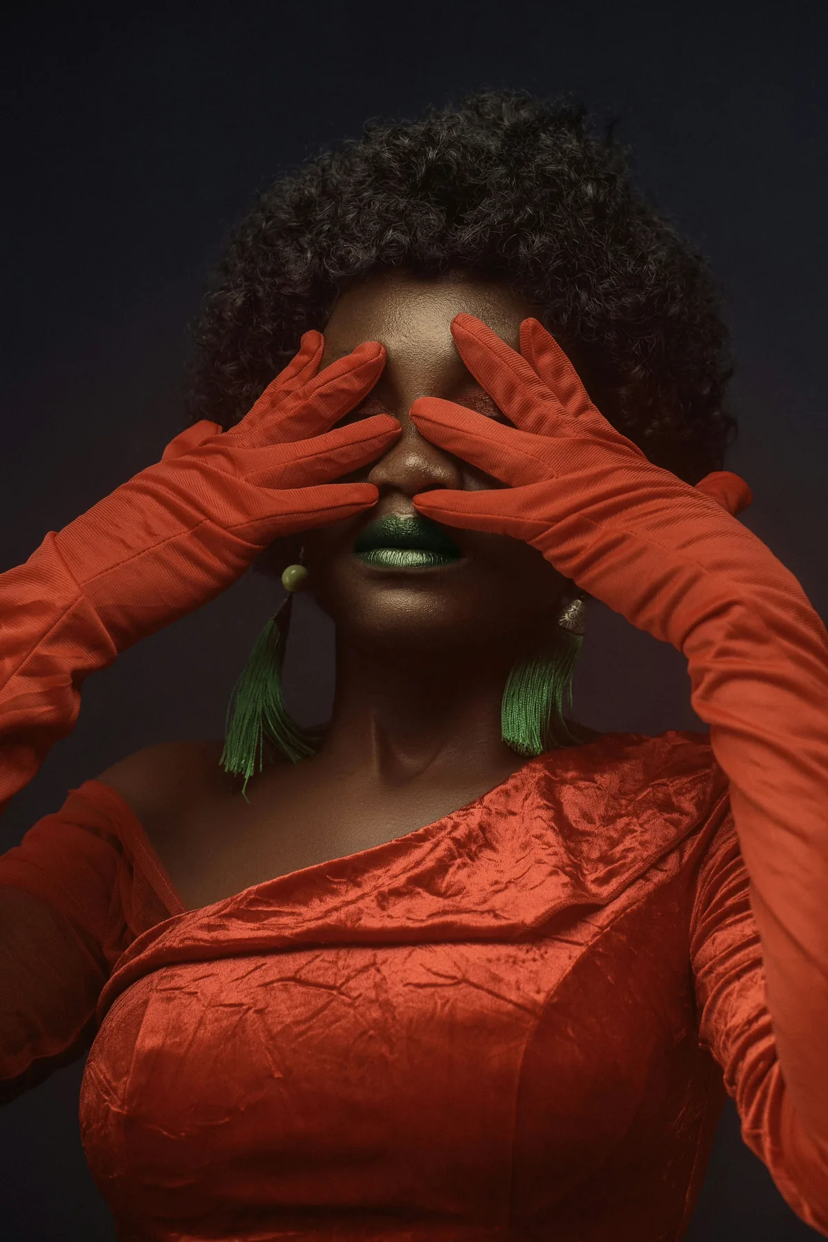 A studio portrait in red and green: gloved hands cover the eyes, lips painted metallic green.