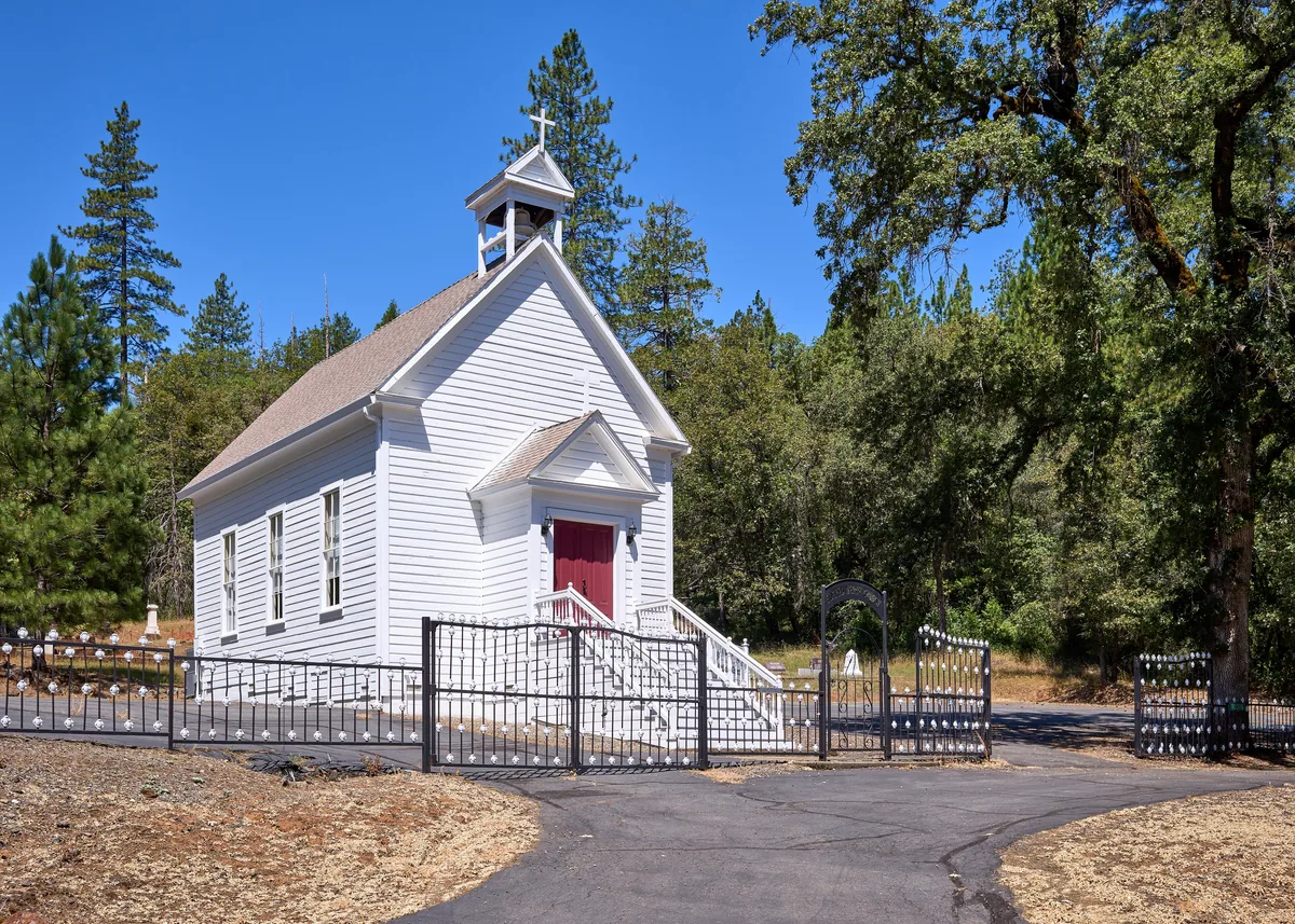 A small white clapboard mission church with a bell cupola among pines and oaks.