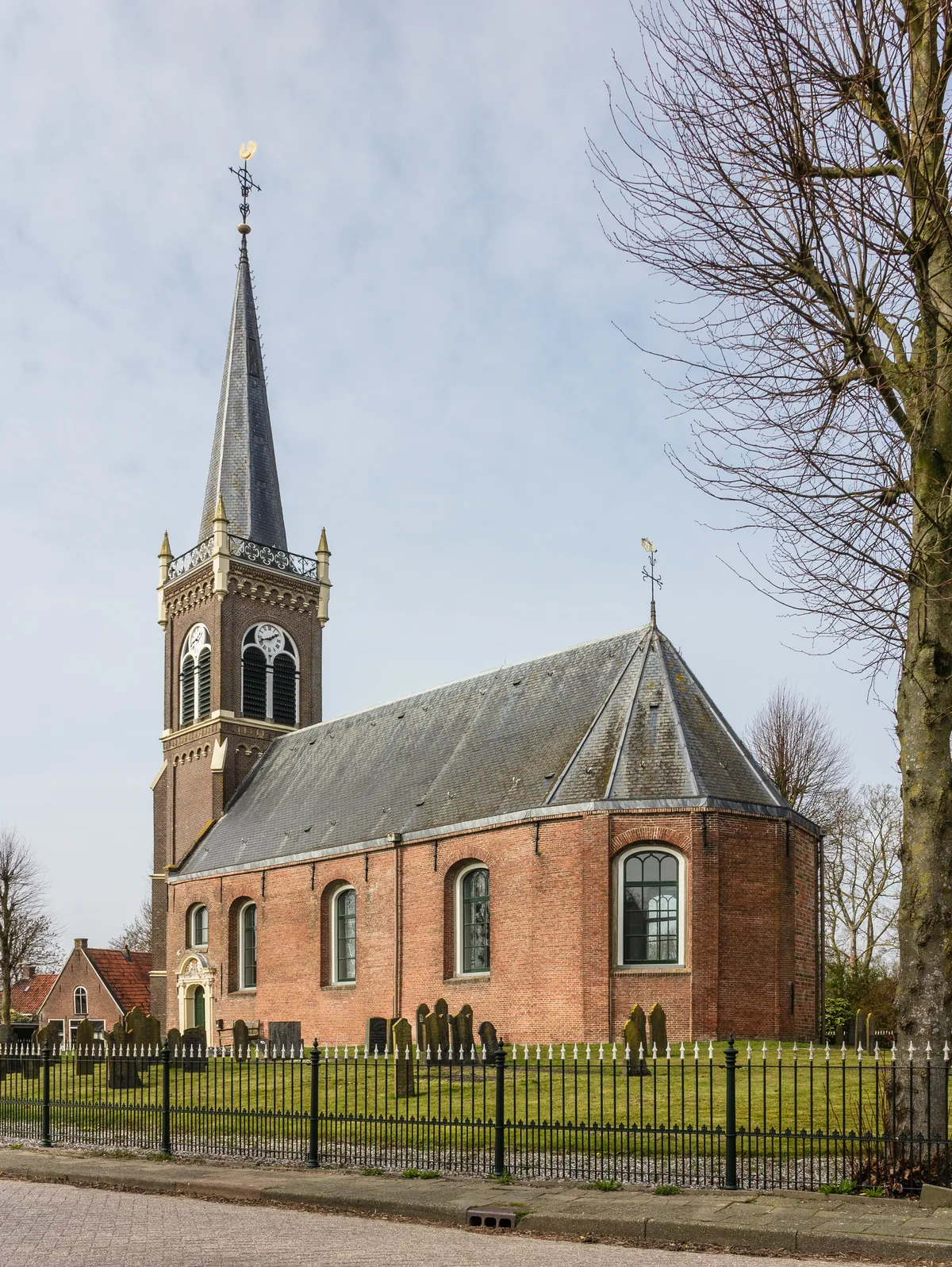A brick village church with a slender spire in Friesland, bare trees and a churchyard in early spring.