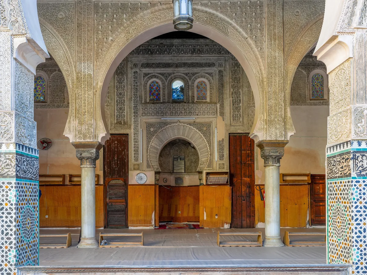 The prayer hall of the Bou Inania madrasa: carved stucco arches, cedar doors and zellige tile around the mihrab.