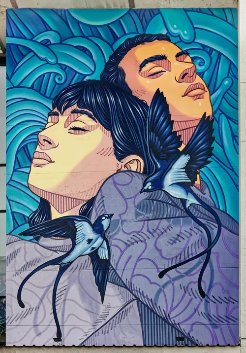 A mural of two faces, eyes closed, wrapped in blue waves and swallows, on a school wall.