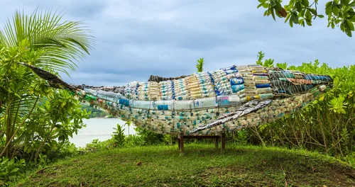 A life-size whale built from collected plastic waste, lying on grass under palm leaves.