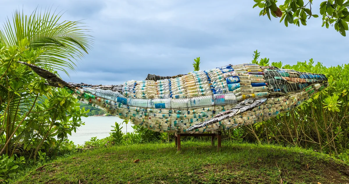 A life-size whale built from collected plastic waste, lying on grass under palm leaves.
