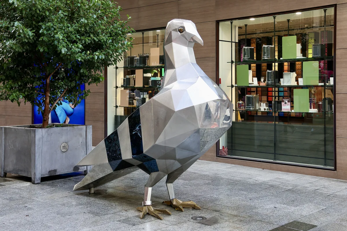An outsize faceted steel pigeon standing on the pavement in front of a shop window.