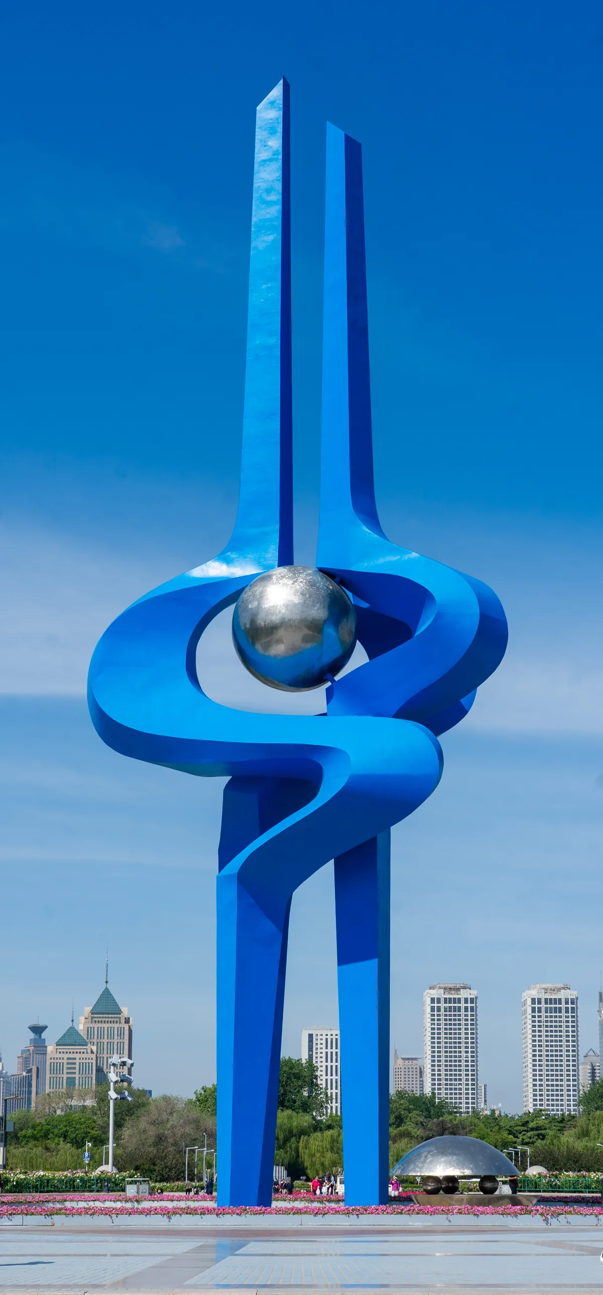 A tall blue sculpture of twisting ribbons and a silver sphere against the sky and the city of Springs.