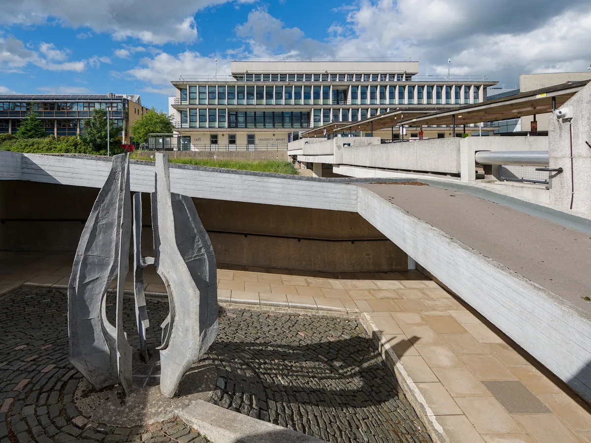 An abstract aluminium sculpture on a concrete terrace in front of the university library.