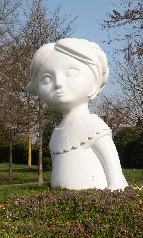 A large white head of a girl with a flower in her hair, standing in a planted verge.