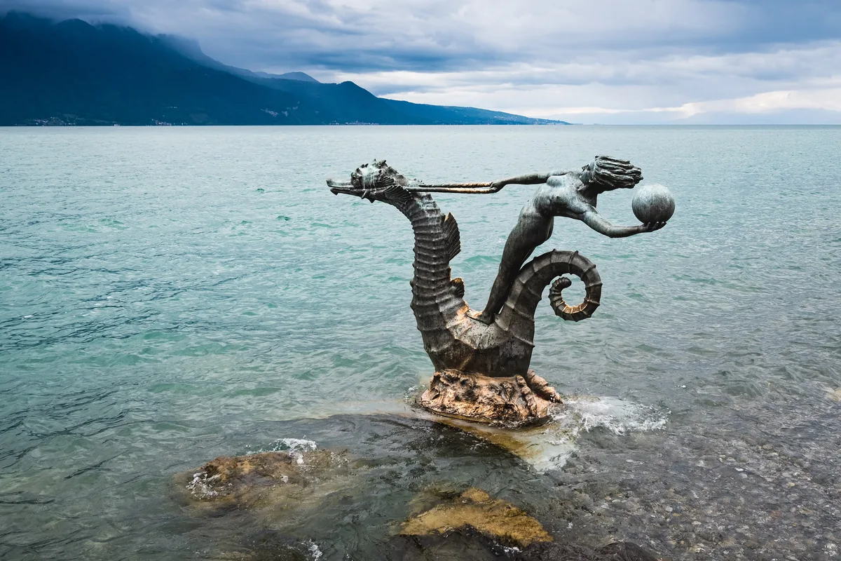A bronze girl riding a seahorse on the rocks of Lake Geneva, the mountains behind her.